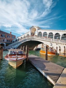 A stunning view of the Rialto Bridge in Venice with wooden gondolas docked on the canal beneath a bright blue sky.