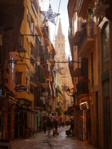 A narrow, lively street in a European city with tall buildings, balconies, shops, and festive decorations hanging overhead, leading towards a historic church steeple in the background.